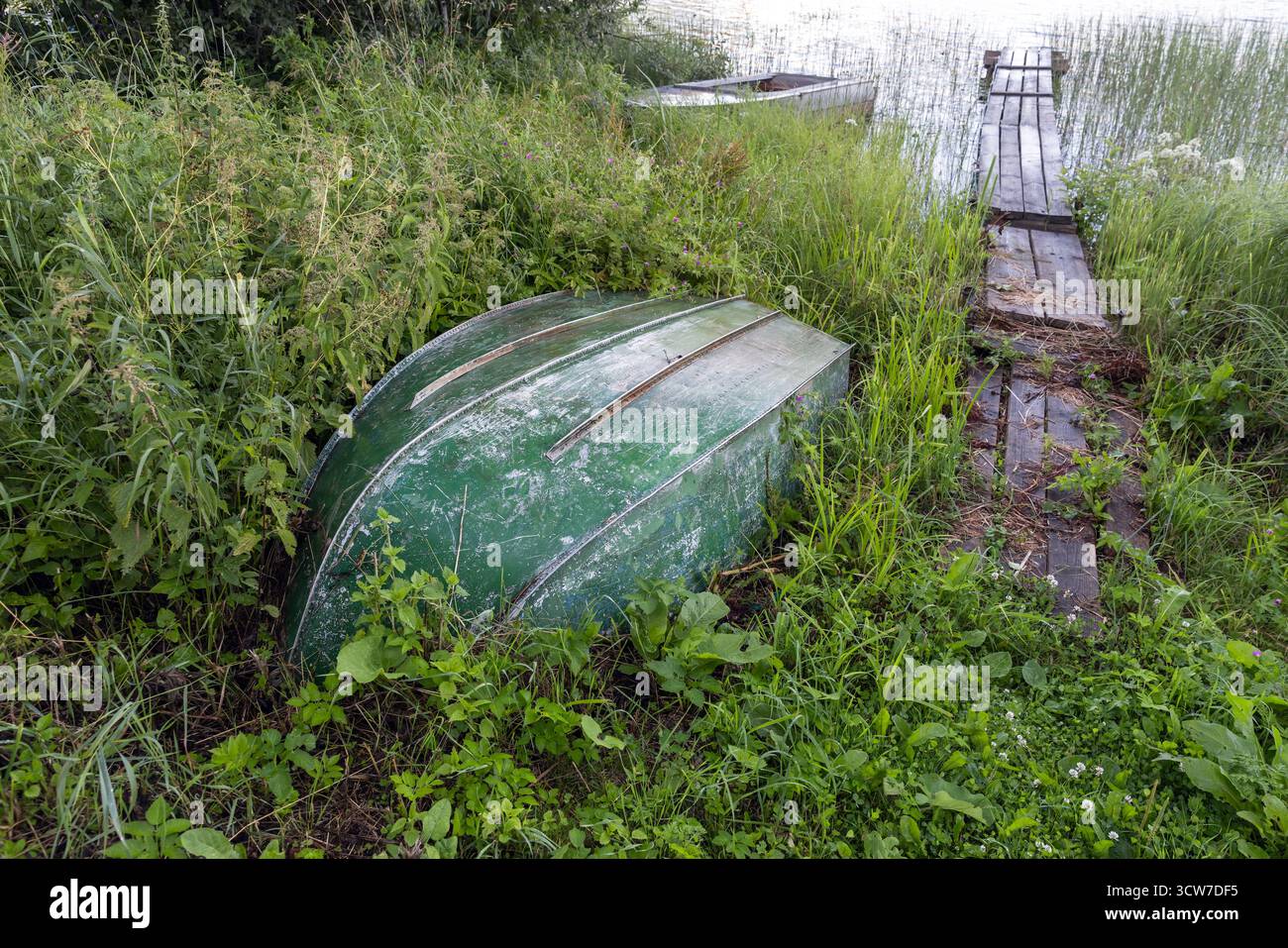 Un bateau vert abandonné se trouve à l'envers dans de hautes herbes à côté d'un quai en bois au bord d'un lac tranquille. Des plantes envahies l'entourent, créant une forge rustique Banque D'Images