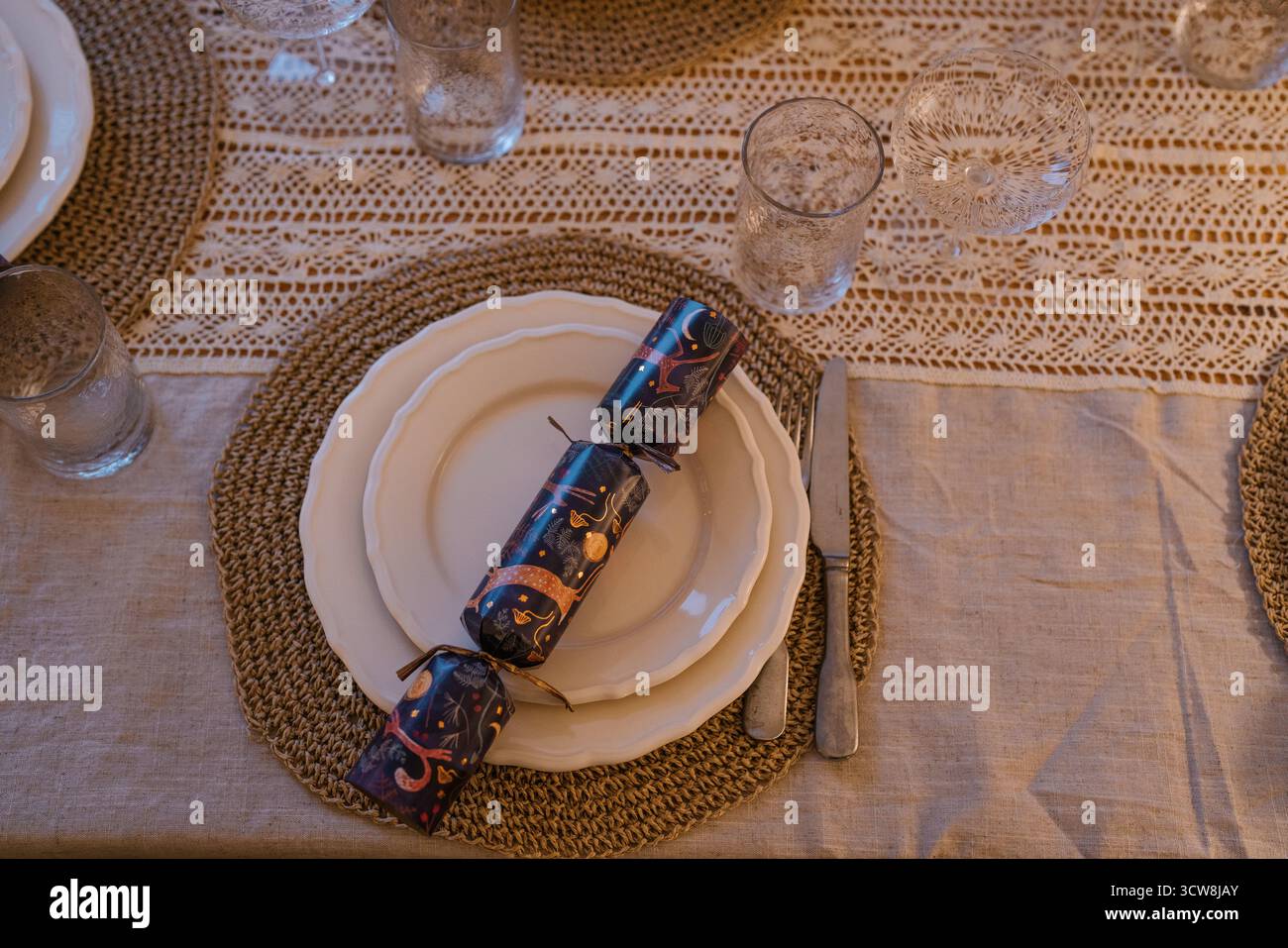 Élégante table de Noël avec assiettes blanches, sets de table tissés, verres en cristal et craquelins festifs sur une nappe en lin avec détails crochet. Banque D'Images