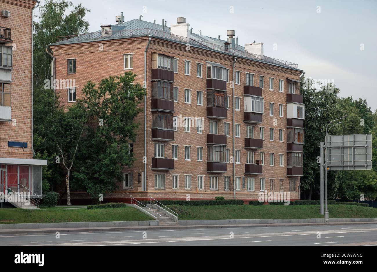 Immeuble résidentiel en briques de cinq étages avec plusieurs balcons, toit plat et cheminées. Les arbres et la verdure sont visibles au premier plan et dans le ciel Banque D'Images