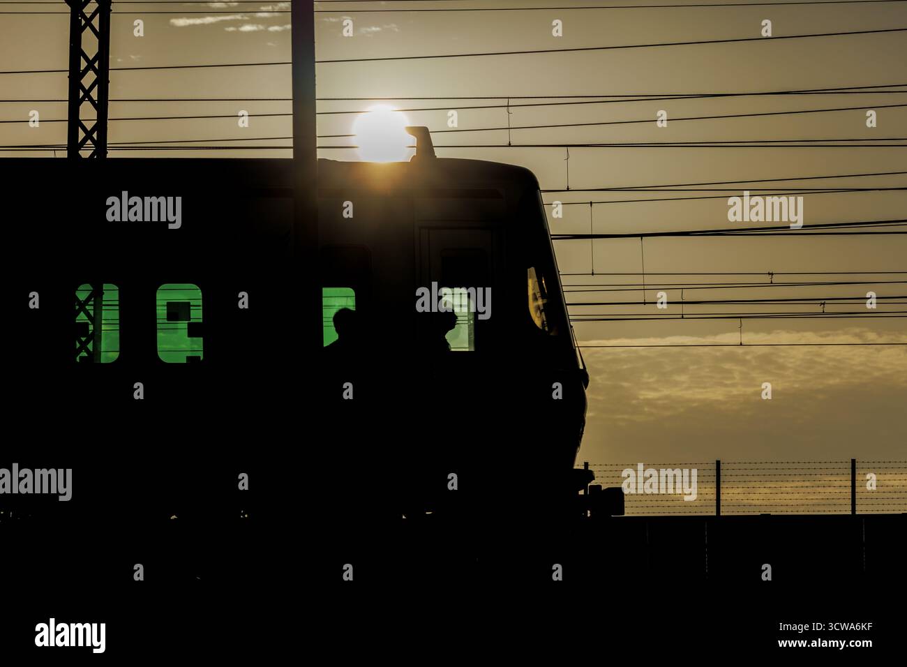 Contours du conducteur et des passagers visibles dans les vitres teintées vertes de la voiture avant du train de banlieue délimitée par une intense lumière dorée du soleil. Banque D'Images