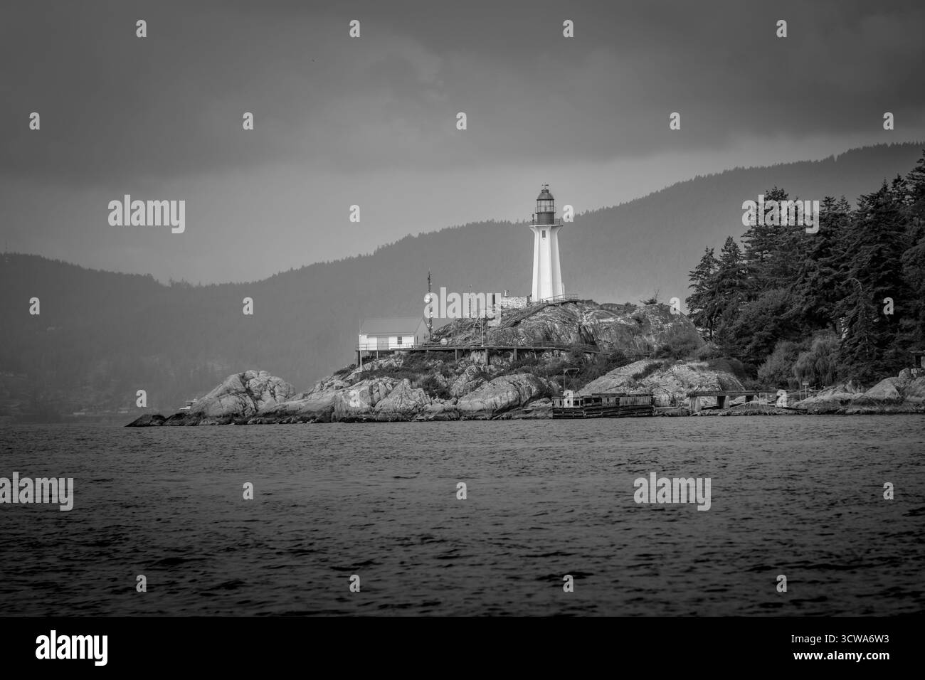 Un phare se dresse sur une île rocheuse alors que des nuages sombres surgissent au-dessus de nous. La mer environnante reflète les tons blancs et gris du ciel, créant une humeur changeante Banque D'Images