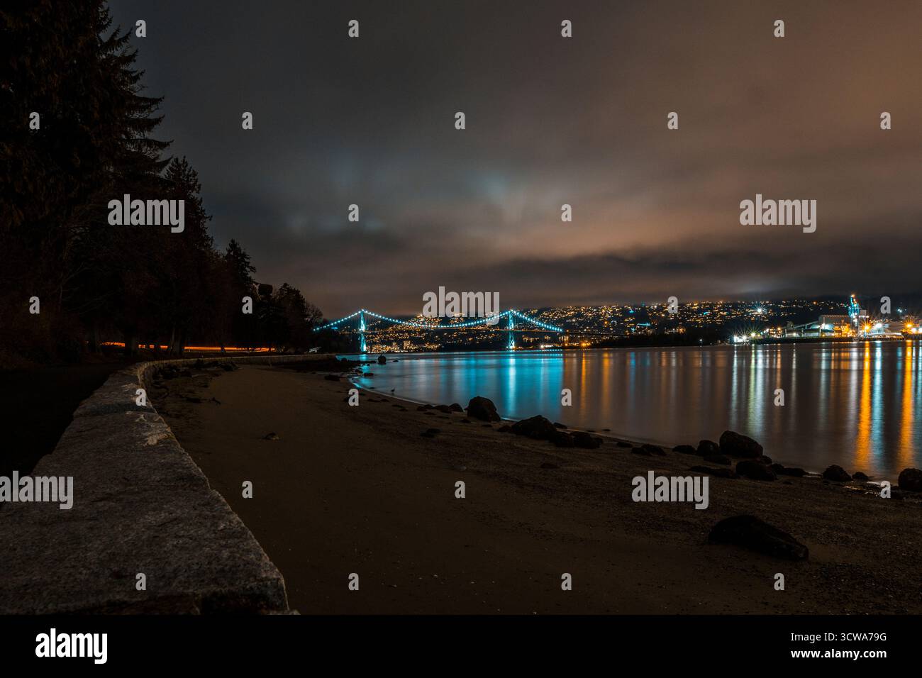 Une scène sereine de front de mer montre les lumières de la ville se reflétant sur l'eau calme la nuit. Des nuages mous dérivent au-dessus, et un pont relie les deux rives. Banque D'Images