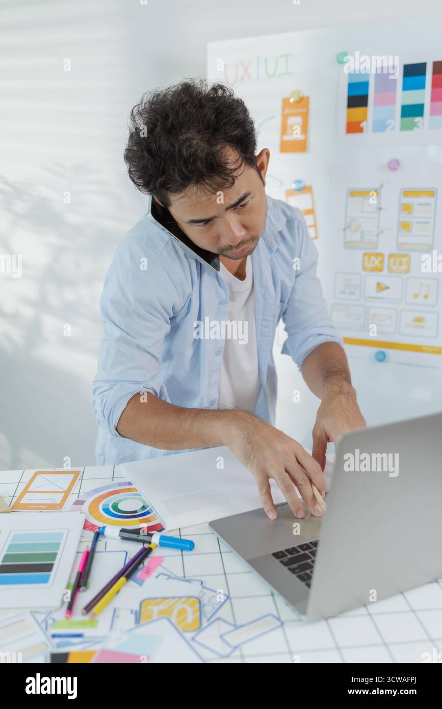 Un concepteur créatif travaille sur la planification UX/UI, analysant des croquis et des prototypes sur un tableau blanc tout en utilisant un ordinateur portable. Le processus montre la pensée de conception Banque D'Images