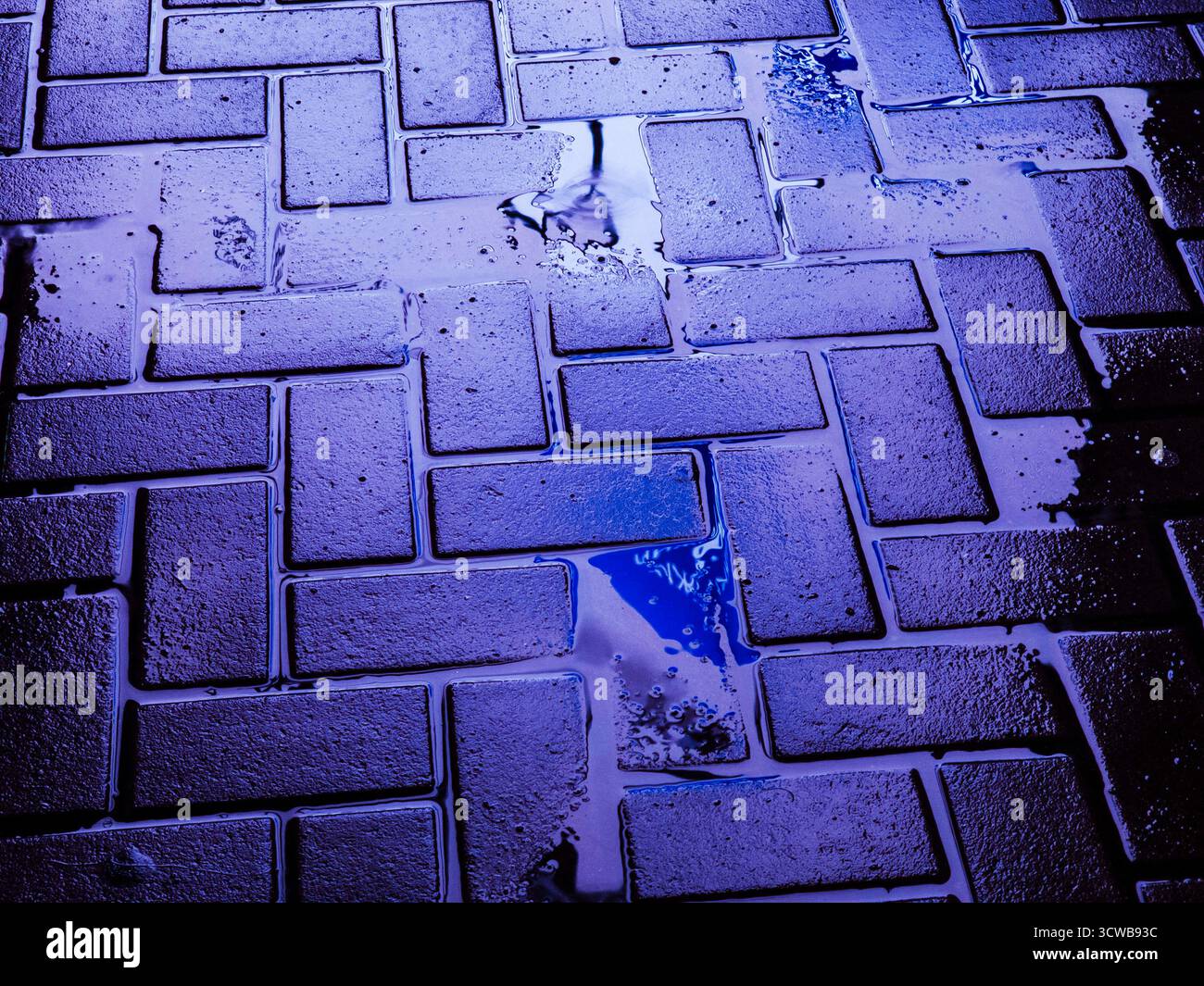 Dalles de pavé urbain mouillées bleu vif et violet, eau accumulée dans des canaux entre des blocs rectangulaires, surface géométrique texturée créant une ambiance Banque D'Images