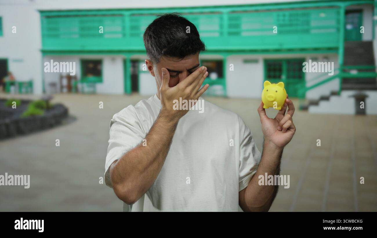 Jeune homme dans une chemise blanche tient une tirelire jaune dans un cadre de ville en plein air, affichant une expression réfléchie sur un fond d'acce vert Banque D'Images