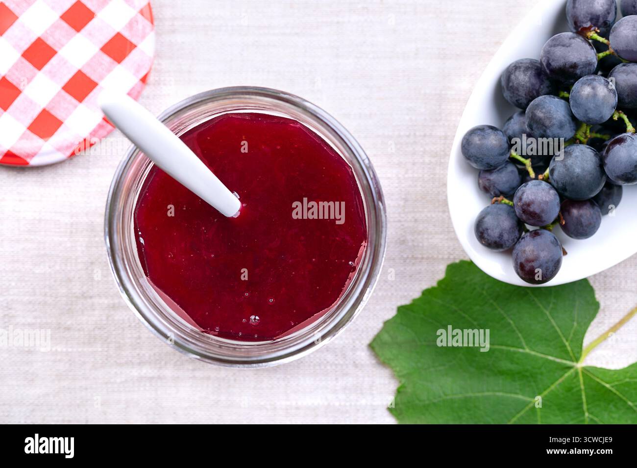 Confiture de raisin faite de raisins bleus, dans un bocal en verre sur lin. Verre ouvert avec bouchon à vis et cuillère en plastique trempé dans de la confiture de raisin maison. Banque D'Images