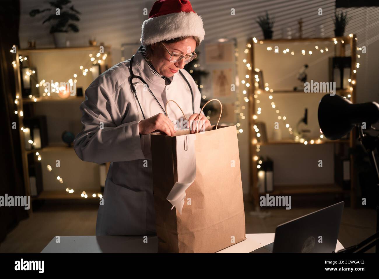 Jeune femme médecin dans des lunettes et chapeau de Père Noël sur le quart déballage de nourriture à emporter dans un sac en papier travaillant la nuit la veille de Noël à l'hôpital. Banque D'Images