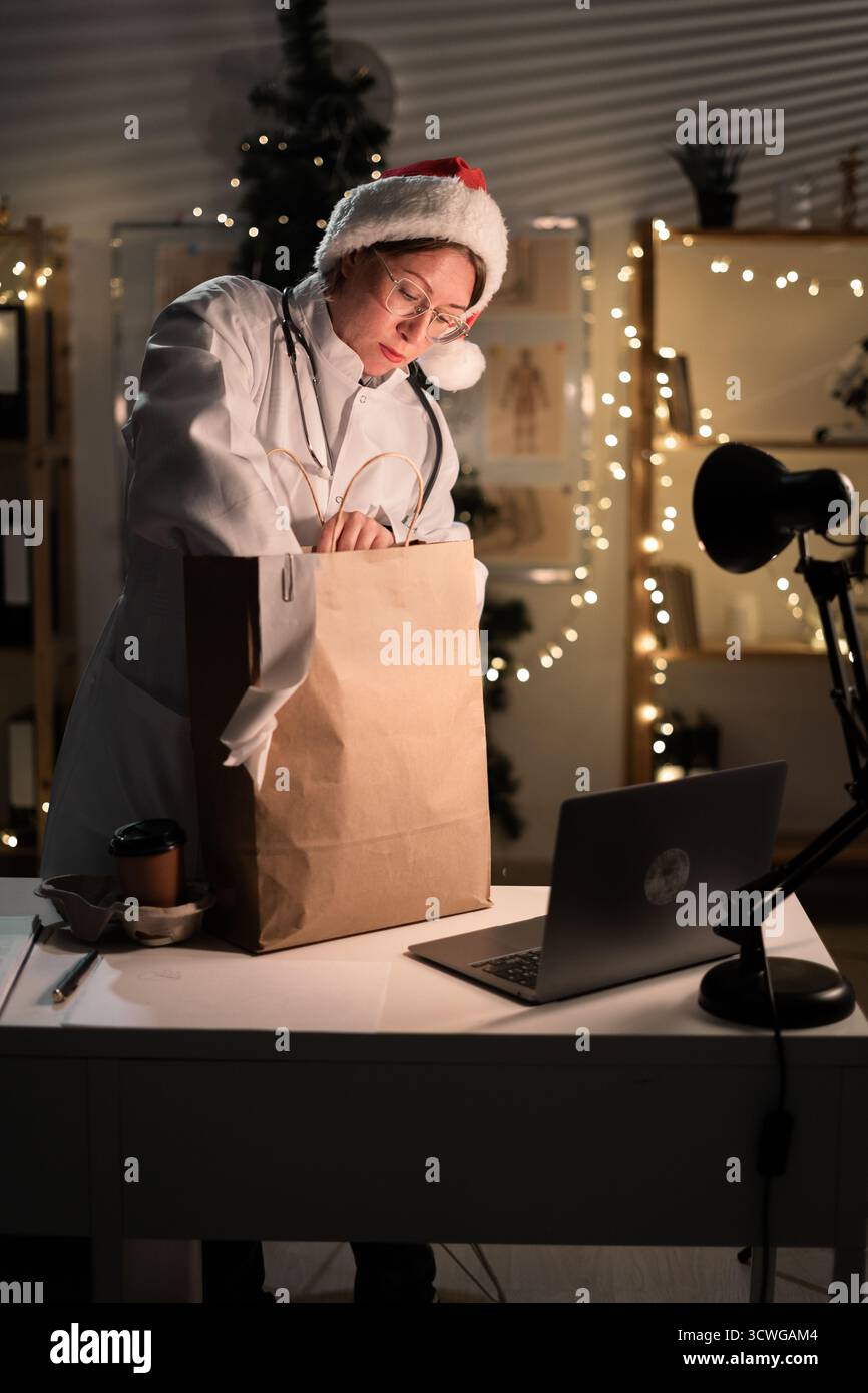 Jeune femme médecin dans des lunettes et chapeau de Père Noël sur le quart déballage de nourriture à emporter dans un sac en papier travaillant la nuit la veille de Noël à l'hôpital. Banque D'Images