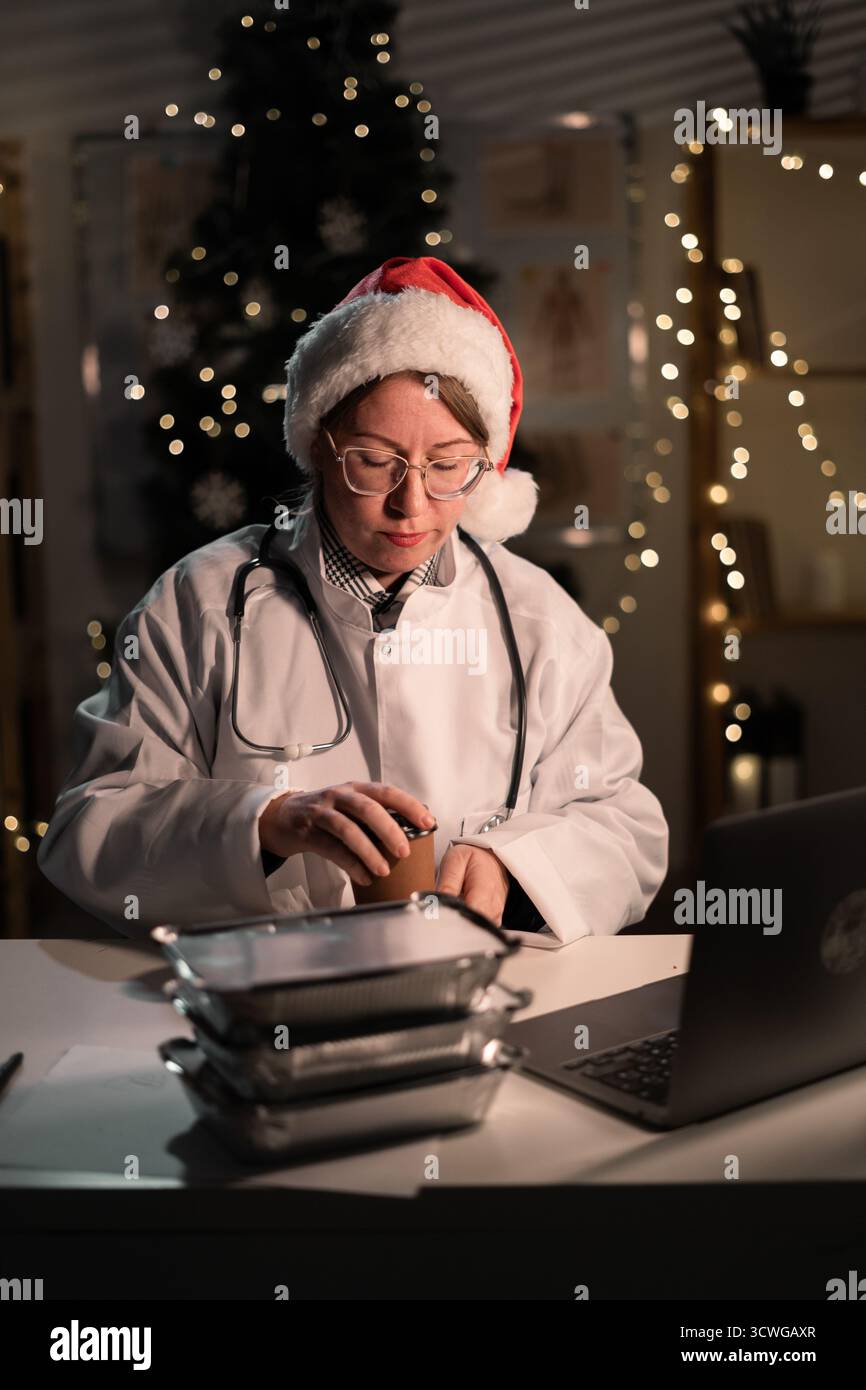 Jeune femme médecin dans des lunettes et chapeau de Père Noël sur le quart déballage de nourriture à emporter travaillant la nuit la veille de Noël à l'hôpital. Vacances, moi Banque D'Images