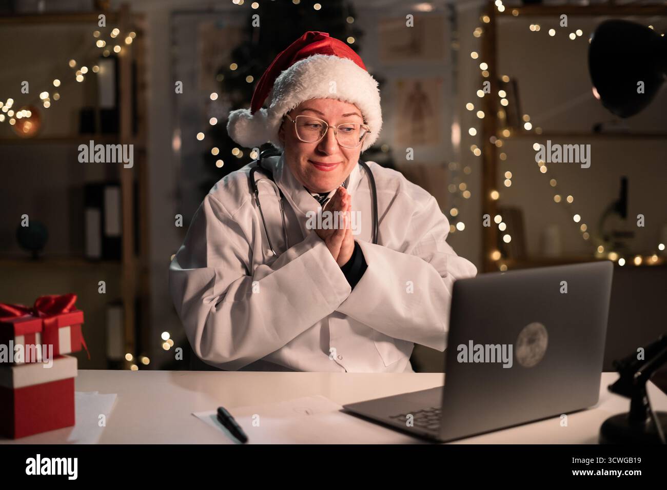 Jeune femme médecin porte chapeau de Père Noël et lunettes à l'aide d'un ordinateur portable tenant les mains dans l'aide-moi s'il vous plaît geste sur le quart de nuit sur Christ Banque D'Images
