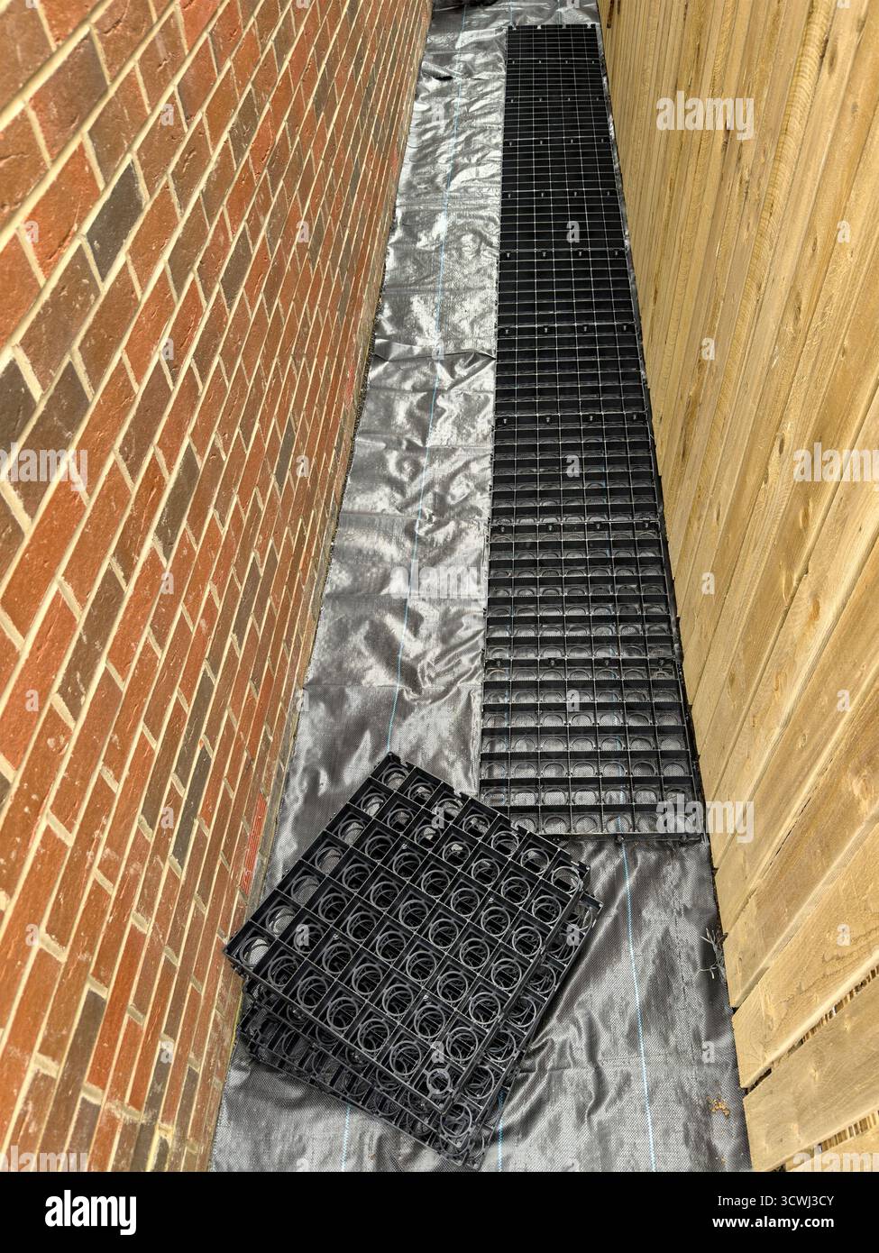 Feuilles de grille en plastique alignées sur un sol recouvert de bâche entre un mur de briques et une clôture en bois dans un espace extérieur étroit - Image de stock capturée avec un smartphone