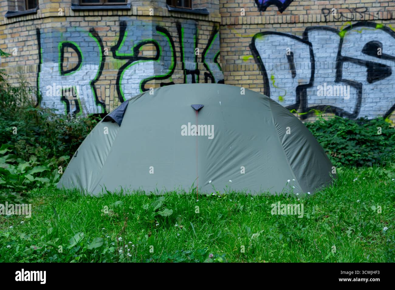Tente sans abri sur herbe près d'un mur de briques avec graffiti à Kreuzberg, Berlin, symbolisant l'inégalité sociale et la vie urbaine Banque D'Images