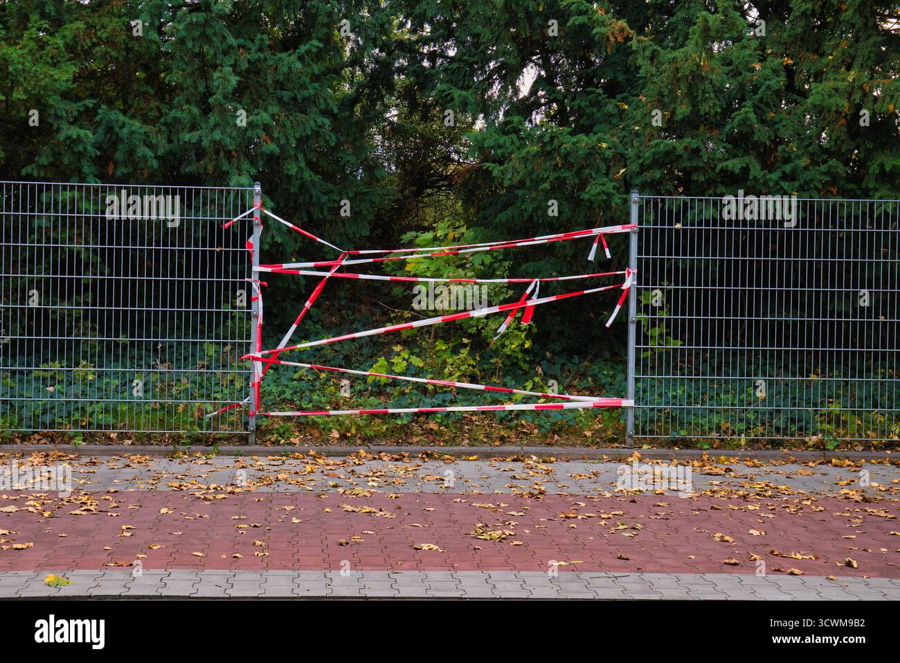 Clôture métallique enveloppée de ruban d'avertissement rouge et blanc bloquant le chemin au milieu des arbres verts et des feuilles d'automne tombées sur un sol pavé Banque D'Images