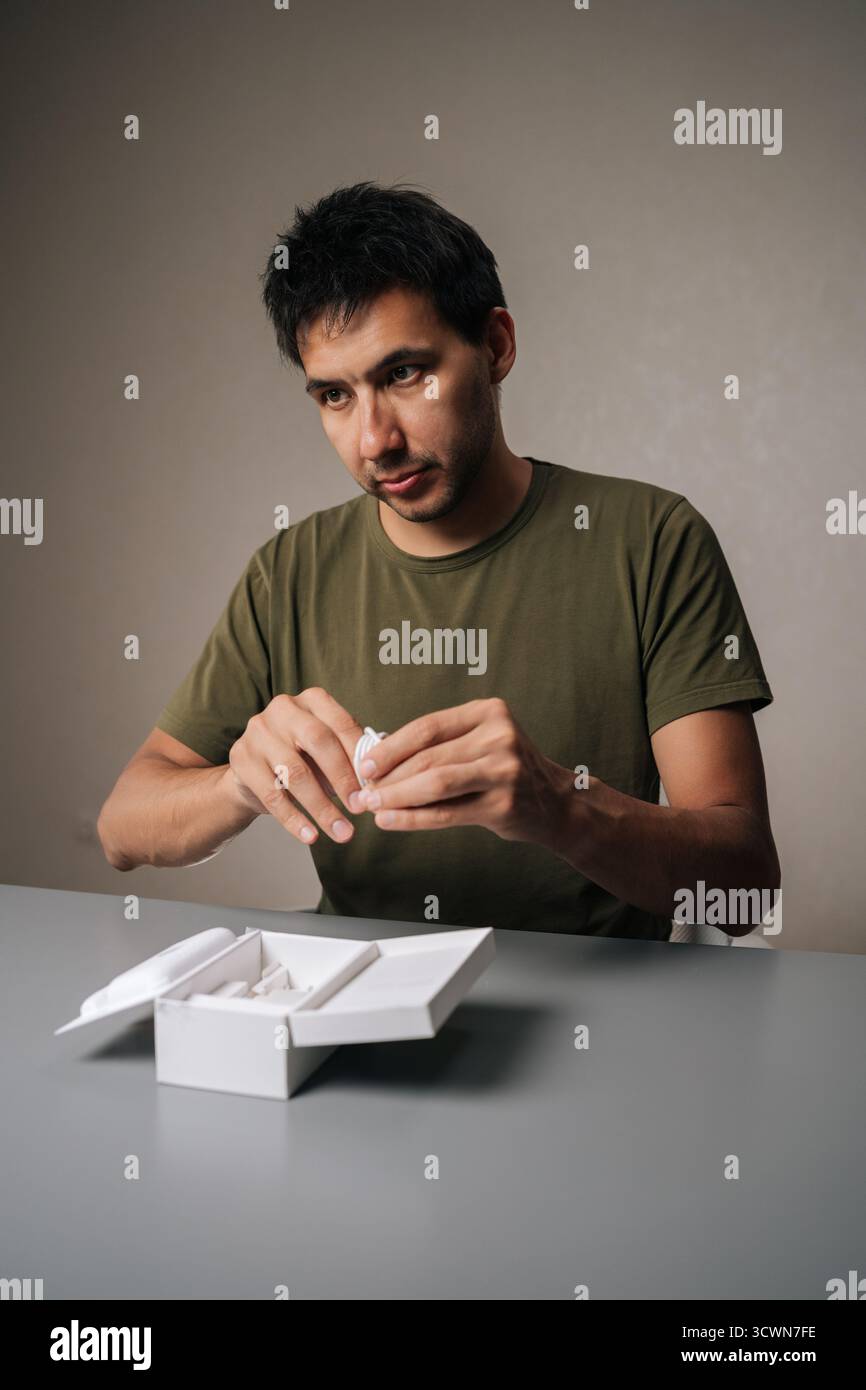 Portrait vertical du client masculin déballant les nouveaux écouteurs sans fil achetés, en retirant de la boîte sur la table grise, exprimant l'excitation Banque D'Images