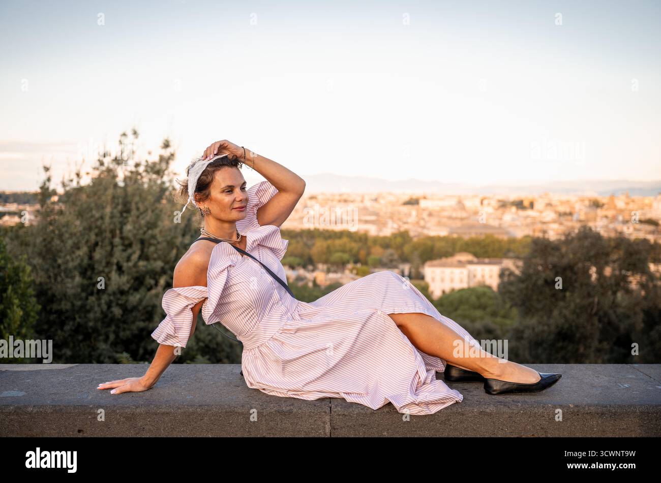 Femme en robe rayée assise sur le mur de pierre au coucher du soleil vue sur la ville Banque D'Images