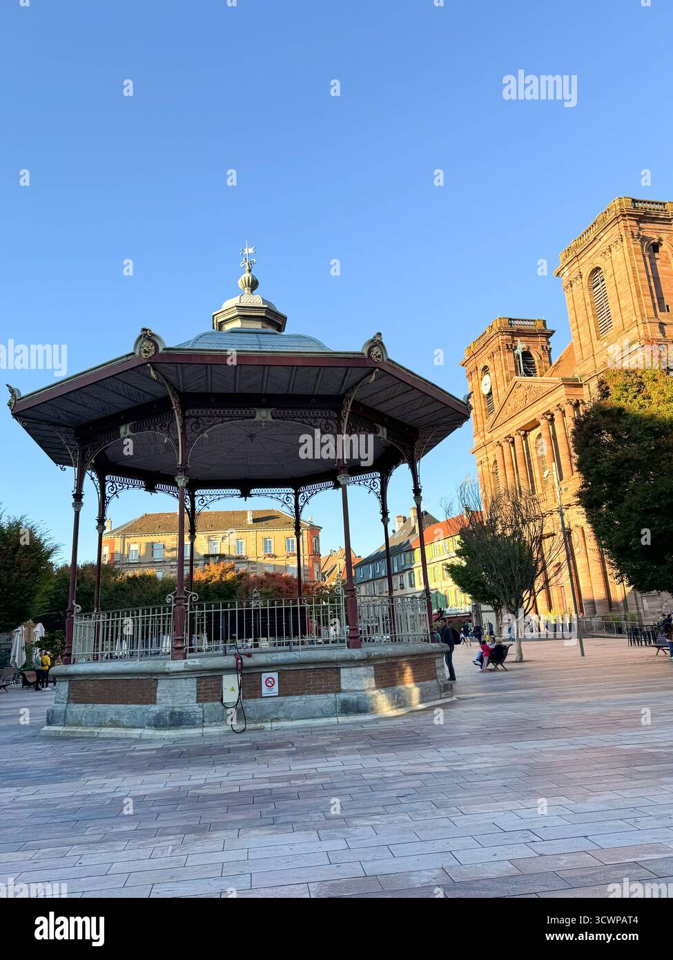 Kiosque à musique orné en fonte sur la place centrale de Belfort, avec église historique et tour de l'horloge en arrière-plan sous la lumière chaude du soleil de l'après-midi - Image de stock capturée avec un smartphone