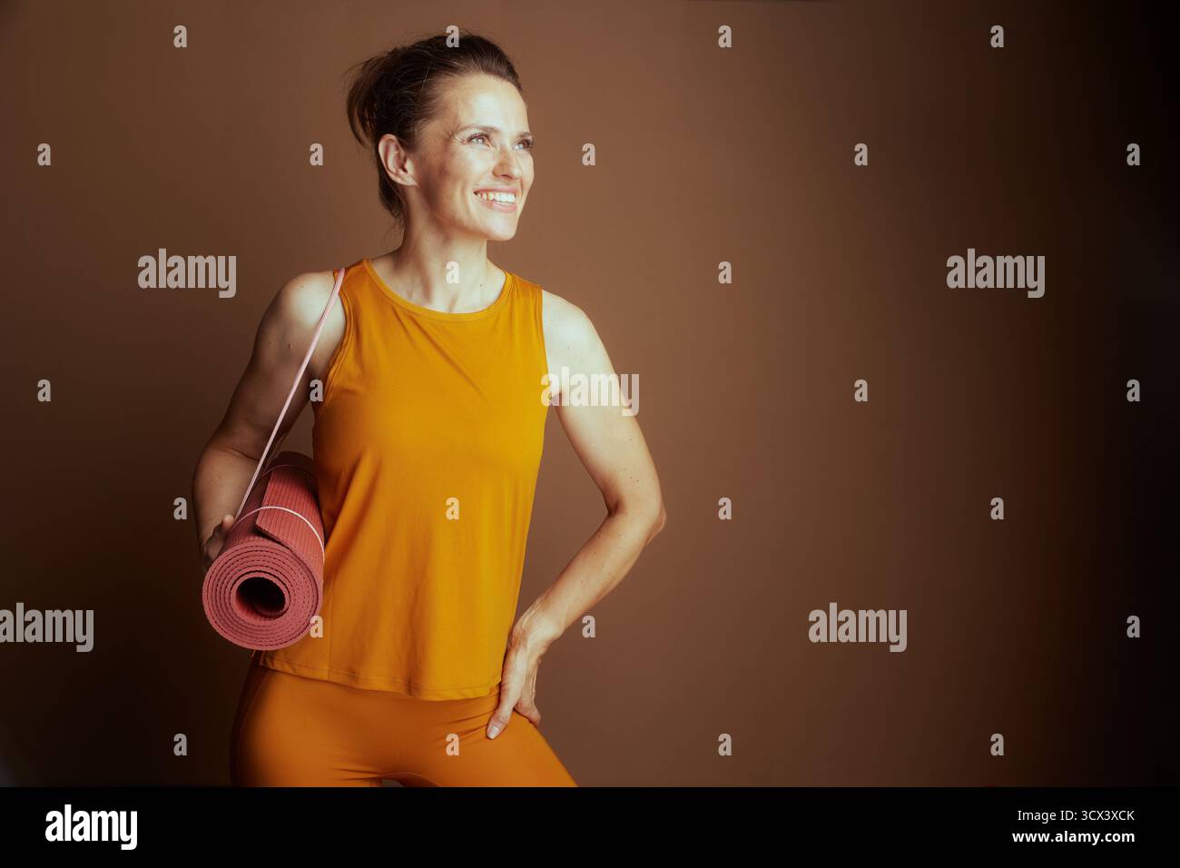 Une femme souriante dans un débardeur orange et des leggings, tenant un tapis de yoga rose, pose avec sa main sur sa hanche, prête pour un entraînement. Elle incarne le fitness Banque D'Images