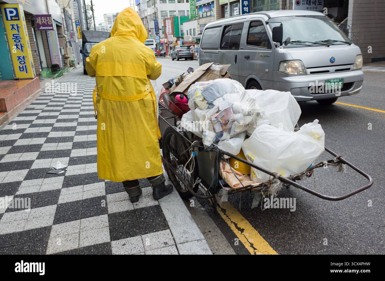 Busan, Corée du Sud - 19 mars 2018 : un travailleur de l'assainissement vêtu d'une combinaison de matières dangereuses jaune vif pousse un chariot lourdement chargé rempli de sacs et de débris Banque D'Images