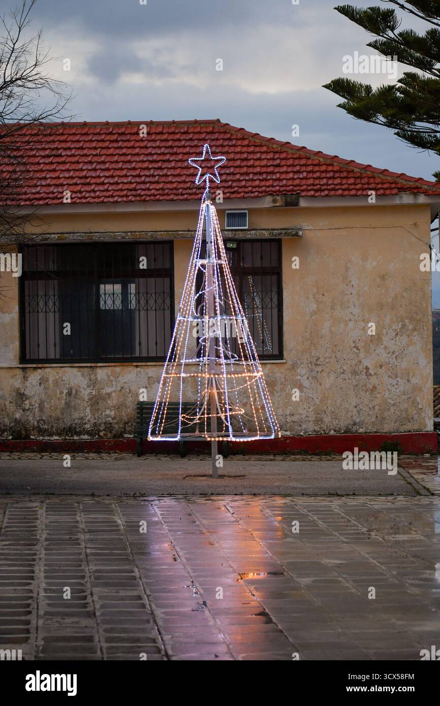 Installation lumineuse moderne d'arbre de Noël brillante sur une place humide dans un village rustique. Une décoration festive de vacances se reflète sur le pavé de pierre humide avec Banque D'Images