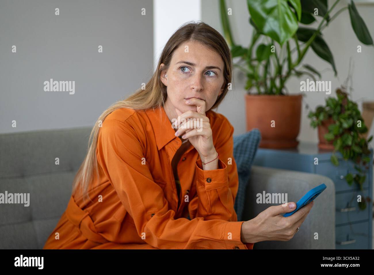 Femme d'âge moyen dans la pensée intense, profonde et anxiété avec smartphone regardant de côté assis sur le canapé Banque D'Images