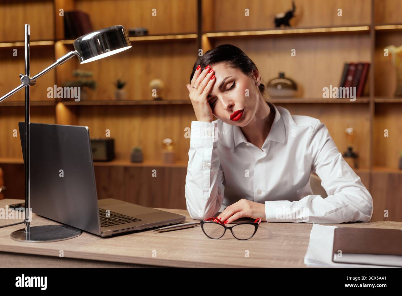 Un employé de bureau fatigué tient la tête à côté d'un ordinateur portable et de lunettes, illustrant la migraine, le stress et la sensibilité aux tempêtes magnétiques sur le lieu de travail. Banque D'Images