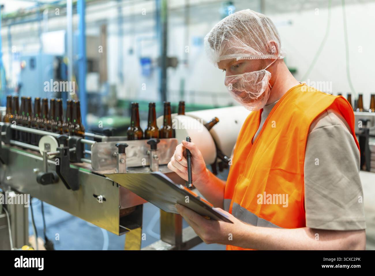 Travailleur dans un filet à cheveux et un masque de visage vérifiant les dossiers de presse-papiers tandis que les bouteilles de bière brune se déplacent le long d'une bande transporteuse de ligne de production dans une usine de boissons Banque D'Images