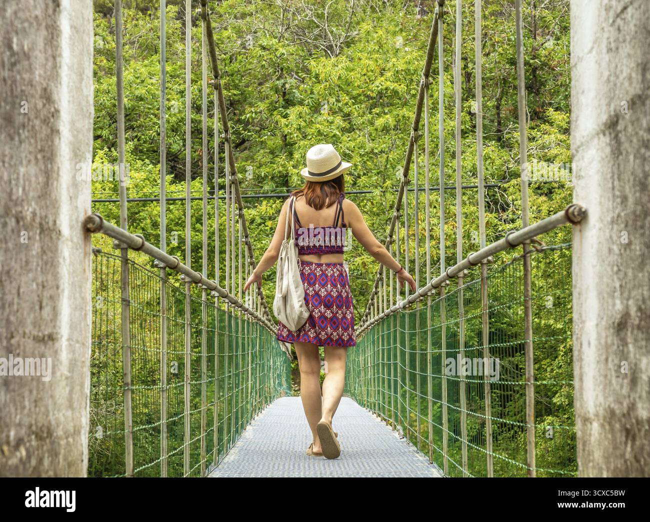 Jeune femme touriste avec un chapeau et un sac marchant sur un pont suspendu au-dessus de la rivière Dobra, profitant d'une aventure estivale pittoresque dans les asturies, en espagne Banque D'Images