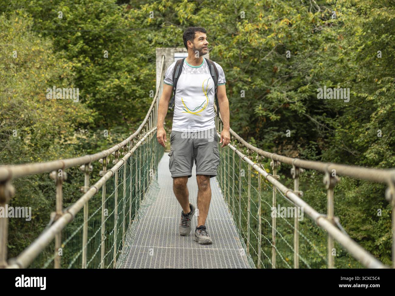 Randonneur masculin avec sac à dos profitant du trekking dans la forêt, marchant sur un pont suspendu métallique au-dessus d'une rivière, entouré d'une végétation luxuriante Banque D'Images