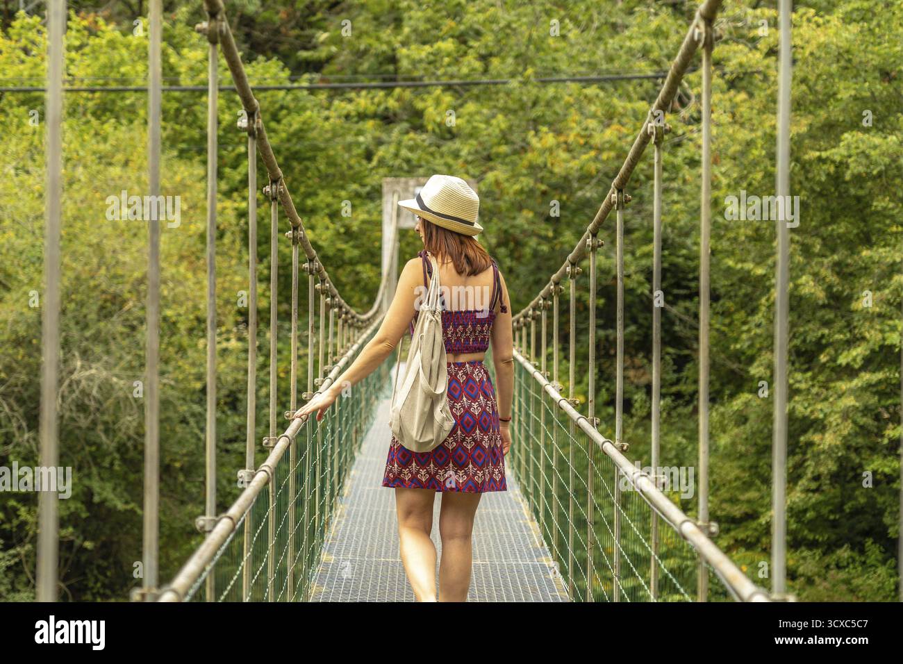 Touriste femme avec chapeau et sac à dos marchant sur un pont suspendu en métal, profitant de la beauté pittoresque d'une forêt verdoyante, expérimentant le frisson Banque D'Images
