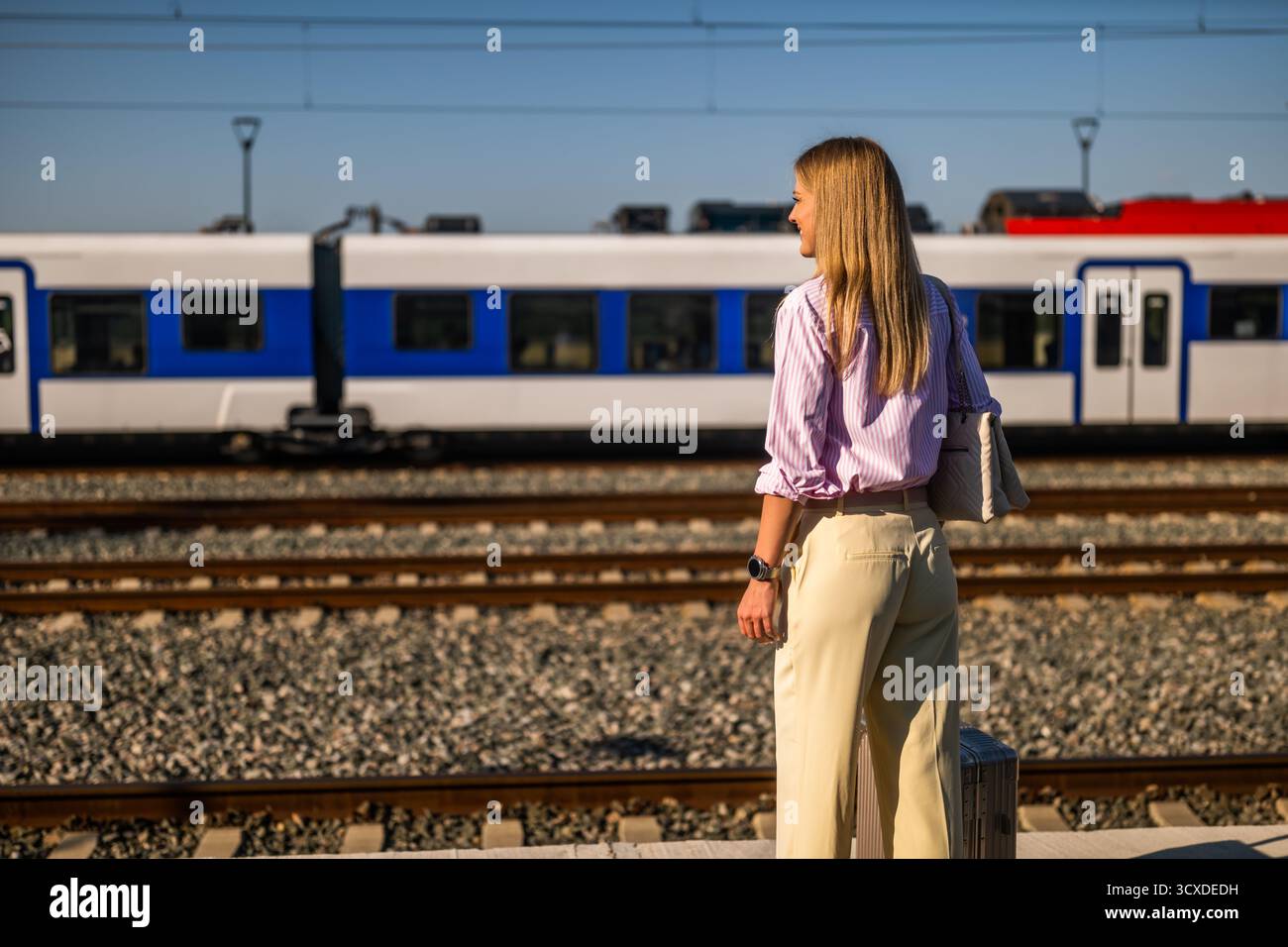 Jeune femme d'affaires debout sur un quai de chemin de fer ensoleillé avec sa valise, attendant son train pendant un voyage d'affaires. Banque D'Images