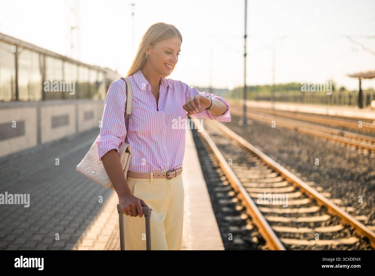 Femme d'affaires confiante et élégante avec valise debout à la gare et regardant sa montre-bracelet. Banque D'Images