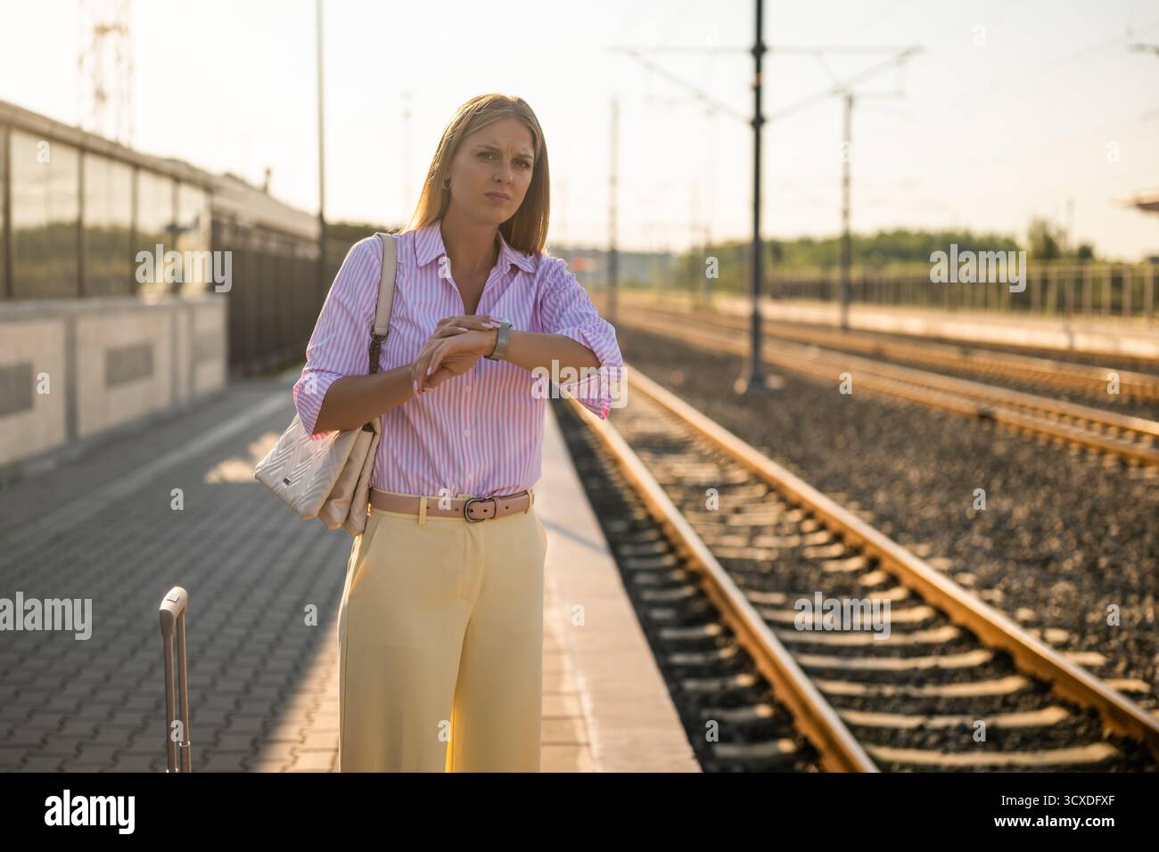 Femme d'affaires mécontente regardant son horloge tout en se tenant debout avec une valise à la gare. Banque D'Images