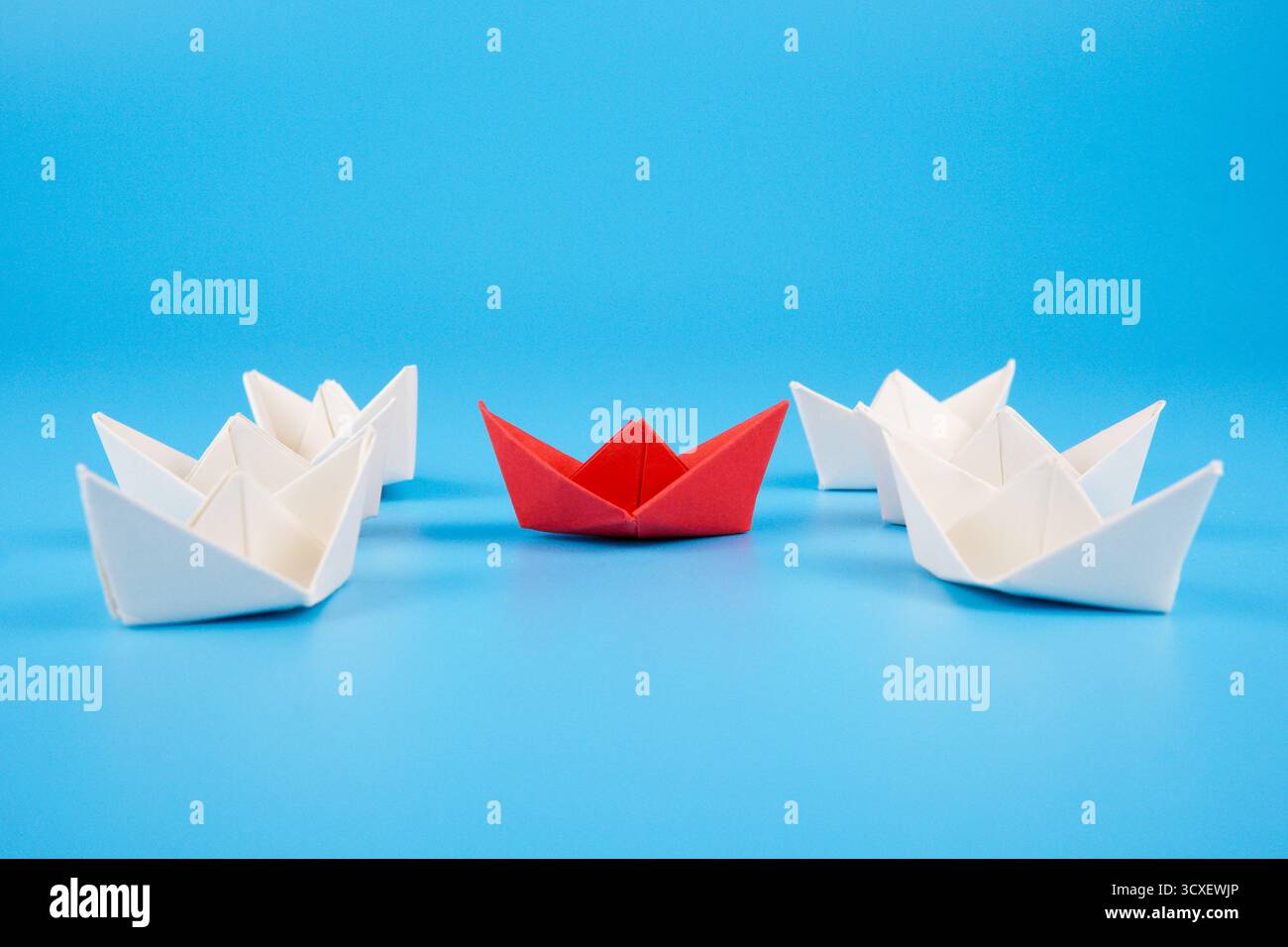 Photo symbolique montrant un bateau de papier rouge entouré de bateaux blancs sur fond bleu, représentant un navire d'aide humanitaire entouré. Banque D'Images