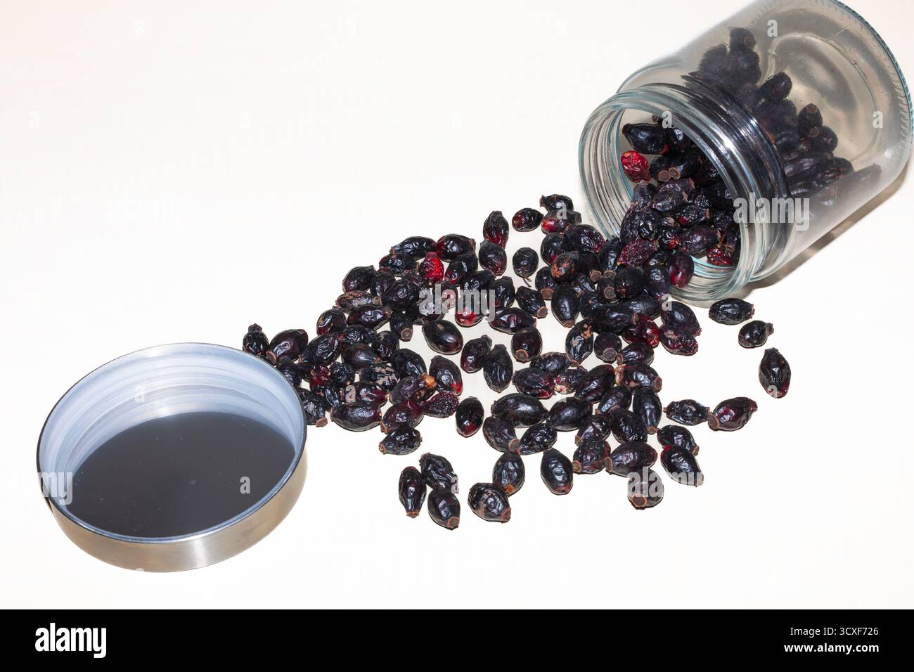 Les hanches de rose séchées (probablement rouge foncé ou presque noires) sont renversées d'un bocal en verre à pointe sur une surface légère, prête pour le brassage ou le stockage. Banque D'Images