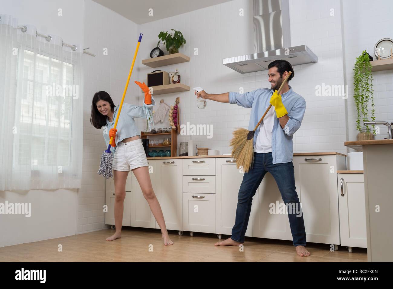 Joyeux couple nettoyant la cuisine ensemble à la maison, en utilisant balai et vadrouille tout en riant et en appréciant les tâches ménagères amusantes, représentant le travail d'équipe, l'amour, Banque D'Images