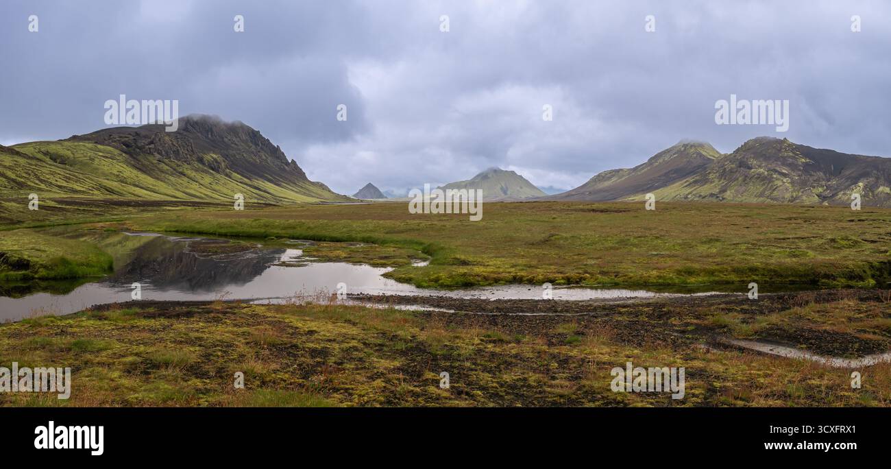 Collines moussues islandaises, étang et montagnes brumeuses sous un ciel nuageux spectaculaire. Banque D'Images