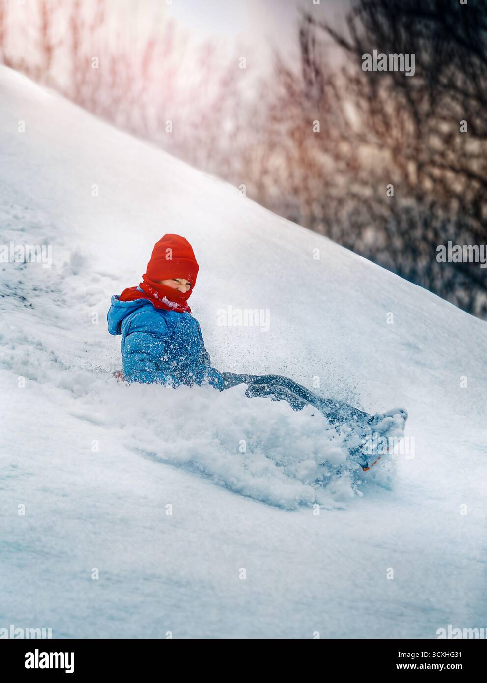 Un enfant vêtu de vêtements d'hiver lumineux glisse sur une colline enneigée, entouré de flocons de neige volants et de lumière du soleil. L'image capture l'excitation de Banque D'Images