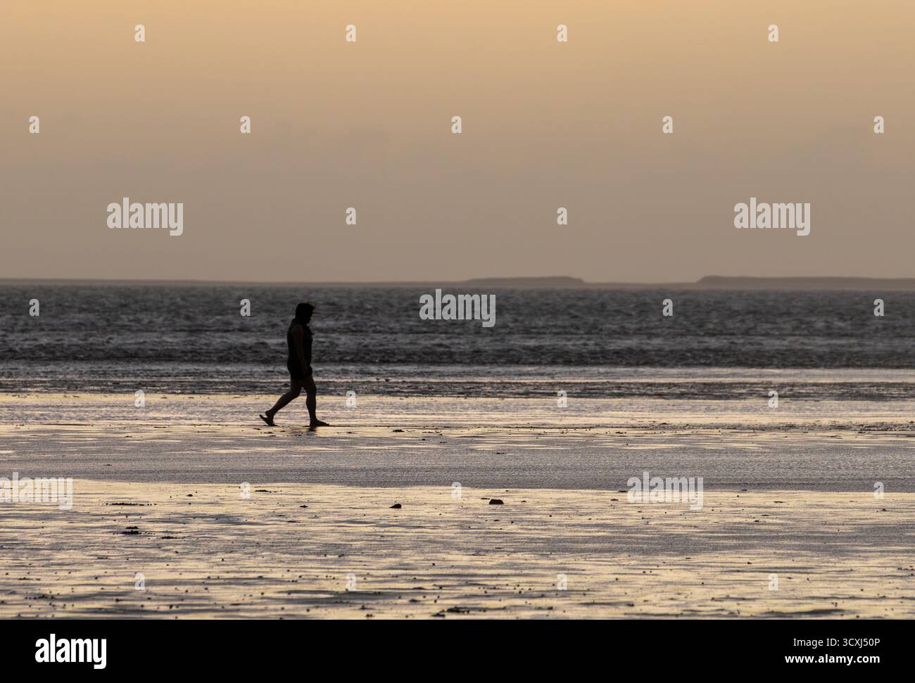 Sunset solitude : Découvrez l'atmosphère paisible comme une personne marche le long du sable humide et réfléchissant pendant l'heure dorée. Un paysage côtier tranquille. Banque D'Images