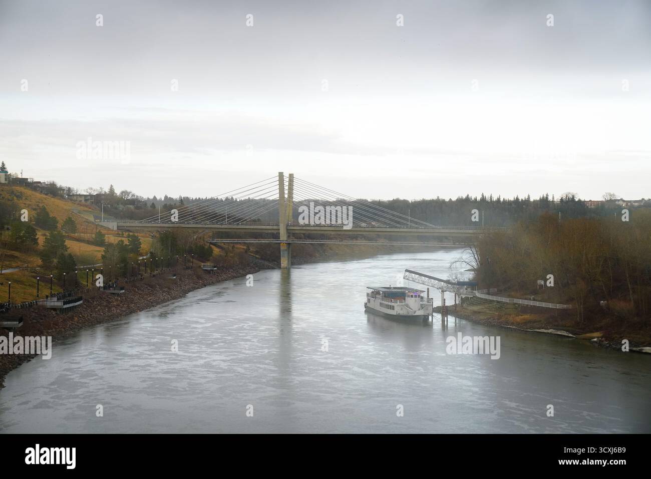 Bateau amarré le long de la rivière Saskatchewan Nord près d'un pont moderne et d'une passerelle en bord de rivière, soulignant un équilibre tranquille Banque D'Images