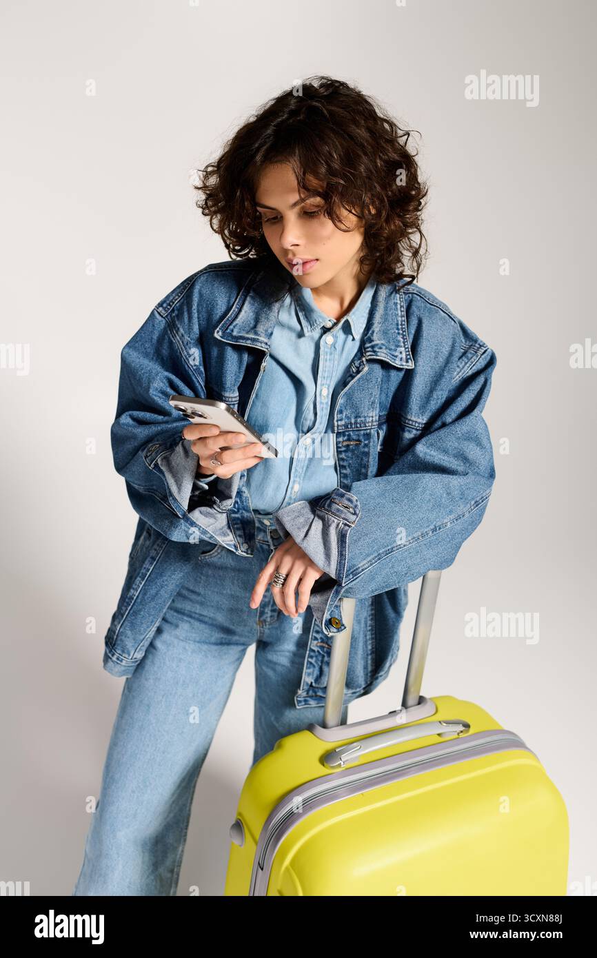 Femme élégante aux cheveux bouclés vérifie son téléphone près d'une valise jaune vif, prête à voyager. Banque D'Images