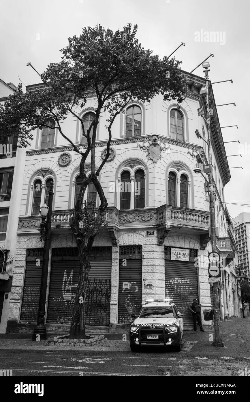 Un policier se tient à côté d'une voiture de patrouille au coin de la rue, surveillant près d'un bâtiment abandonné et arboré et d'un arbre solitaire en noir et blanc Banque D'Images
