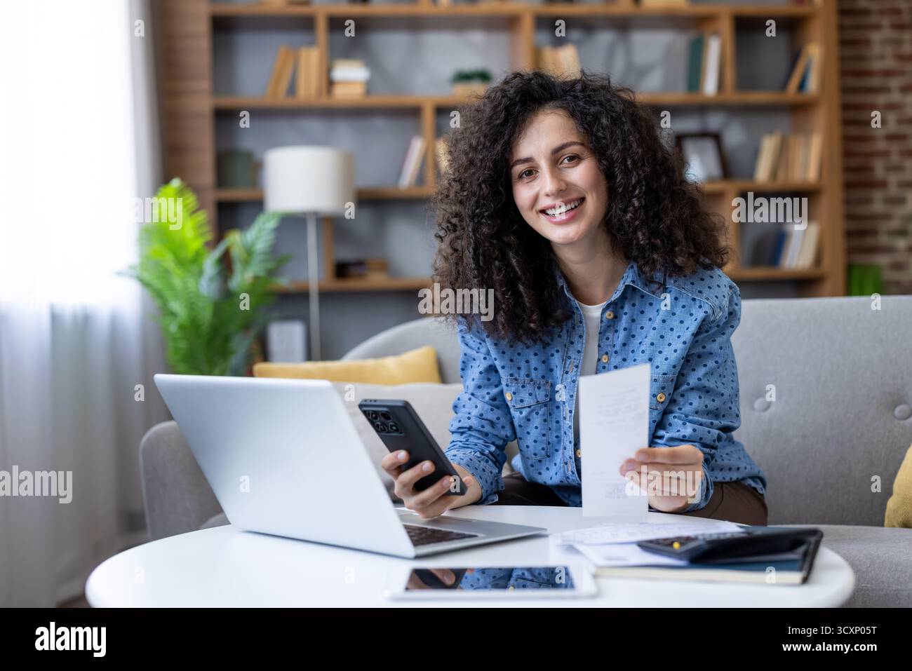 Femme souriante tenant une facture papier, utilisant un smartphone et un ordinateur portable pour gérer son budget personnel et sa planification financière dans un cadre confortable à la maison Banque D'Images