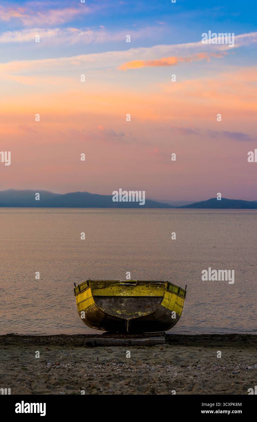 Vieux bateau en bois reposant sur le rivage au coucher du soleil avec des reflets colorés du ciel sur le lac Ohrid et des silhouettes de montagne lointaines Banque D'Images