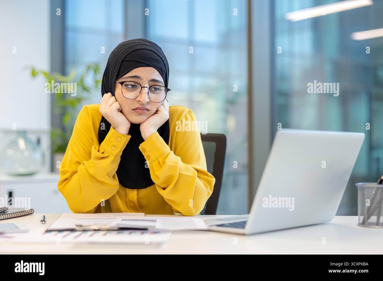 Jeune femme musulmane portant un hijab et des lunettes, posant son visage sur ses mains, exprimant l'ennui, l'épuisement professionnel et le stress tout en travaillant sur un ordinateur portable chez HE Banque D'Images
