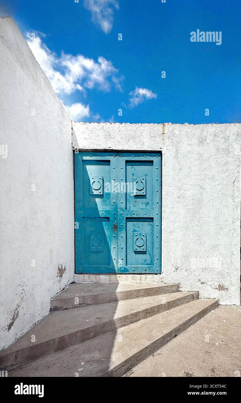 Porte vintage bleue placée contre un mur de stuc blanc avec des marches, sous un ciel bleu vif avec des nuages éparpillés Banque D'Images