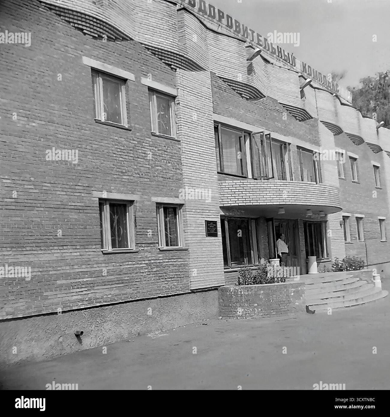 Une photo d'archives des années 1980 fournit une vue détaillée de l'entrée du 'Banno-Ozdorovitelny Kompleks' (complexe de bain et de santé) sur la rue Kommunarov (aujourd'hui rue Torskaya) à Sloviansk. L'image met en valeur l'architecture fonctionnaliste soviétique tardive du bâtiment, avec des briques ornées et une baie d'entrée incurvée. Les bains publics étaient des centres sociaux essentiels en URSS. Il s’agit d’un instantané nostalgique de l’infrastructure urbaine quotidienne et de la vie tranquille d’un Donbass paisible avant la guerre Banque D'Images