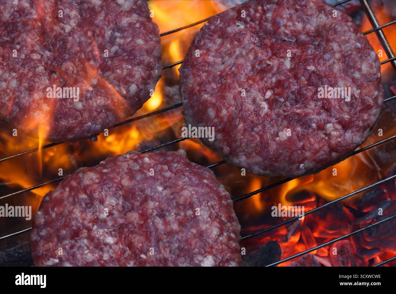 Trois délicieuses galettes de hamburger crues cuites sur un barbecue à charbon chaud avec des flammes qui s'élèvent autour de lui, créant une délicieuse cuisine estivale SC Banque D'Images