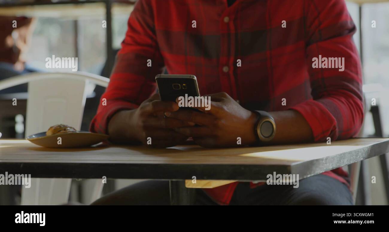 Homme portant une chemise à carreaux rouge et un écran de smartphone défilant smartwatch dans un café, avec une plaque de pâtisserie Banque D'Images