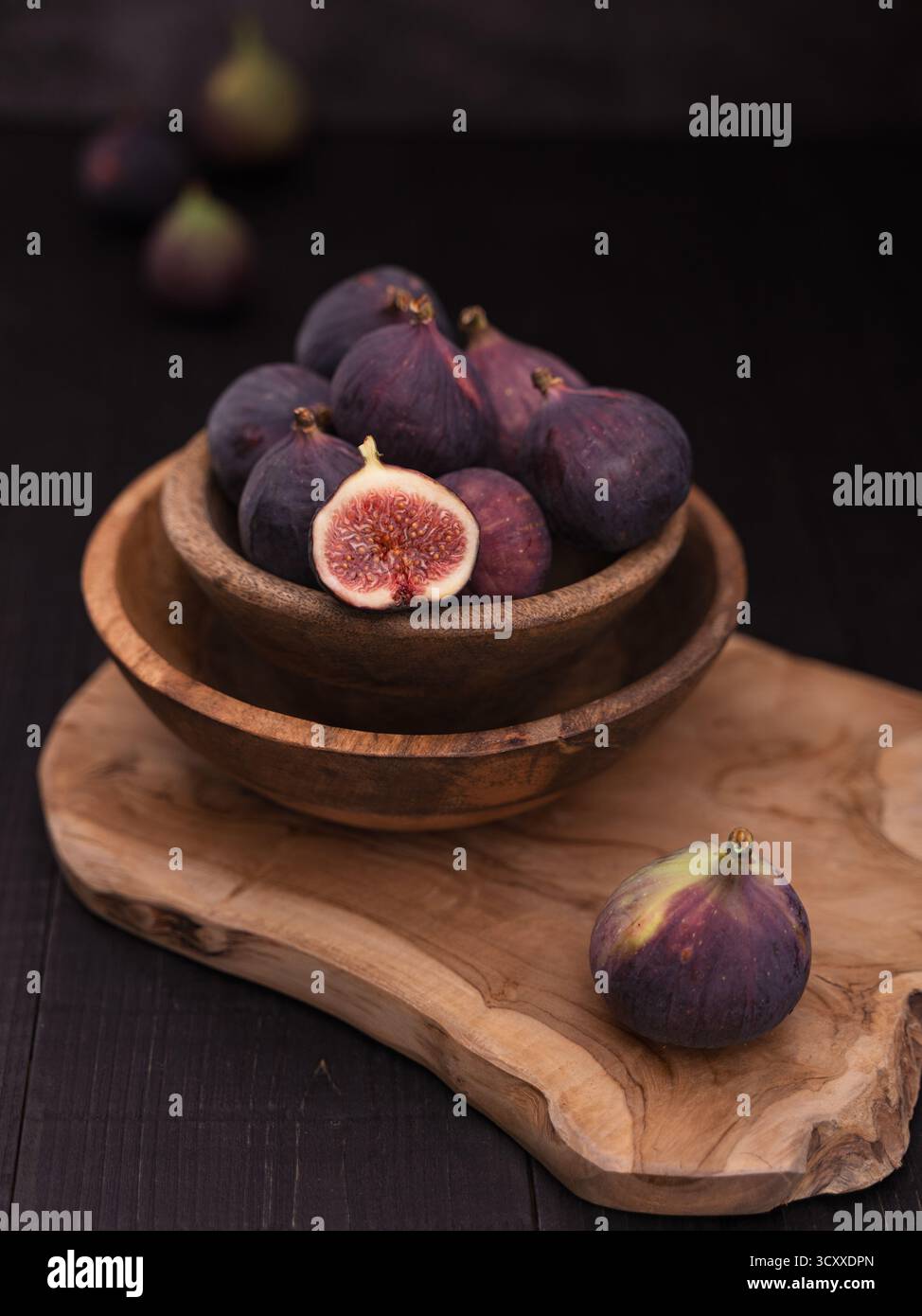 Demi-tranches de figue dans un bol en bois sur une planche de service avec des figues entières à l'avant et à l'arrière, élégante nature morte de nourriture noire Banque D'Images
