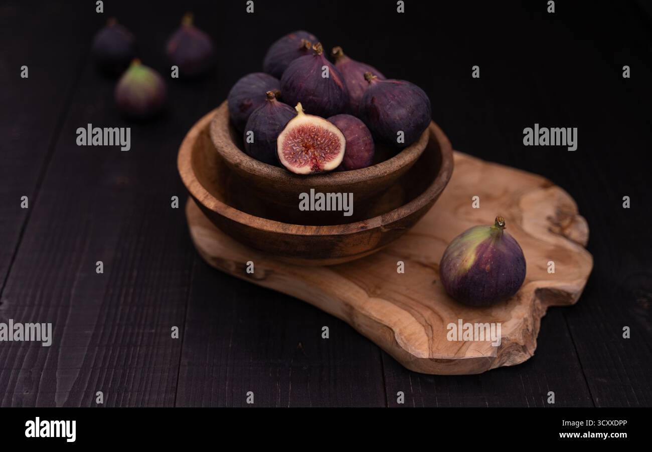 Figue à moitié tranchée dans deux bols en bois empilés sur une assiette de service avec des figues entières disposées à l'avant et à l'arrière, nature morte sombre rustique Banque D'Images